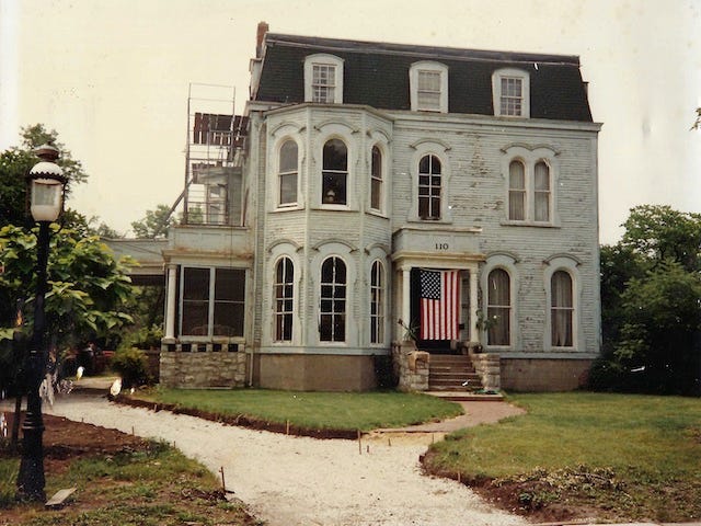 Second Empire Victorian house with green shingle mansard roof and peeling light blue paint. Long gravel driveway is dug up. An antique streetlight is in the foreground.