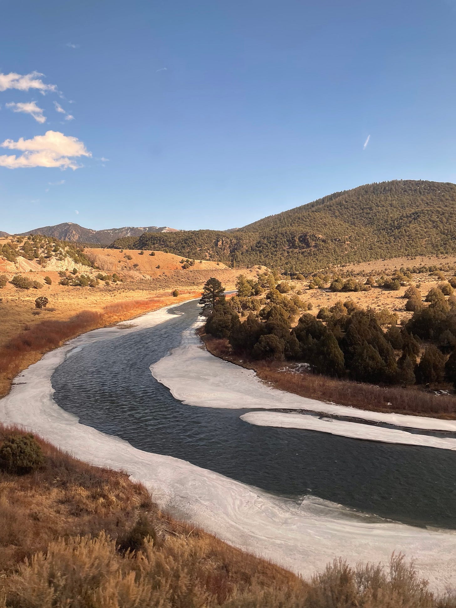 Clear blue skies as a river snakes through a valley, patches of ice visible at the sides. The hills in the distance are covered in green trees.