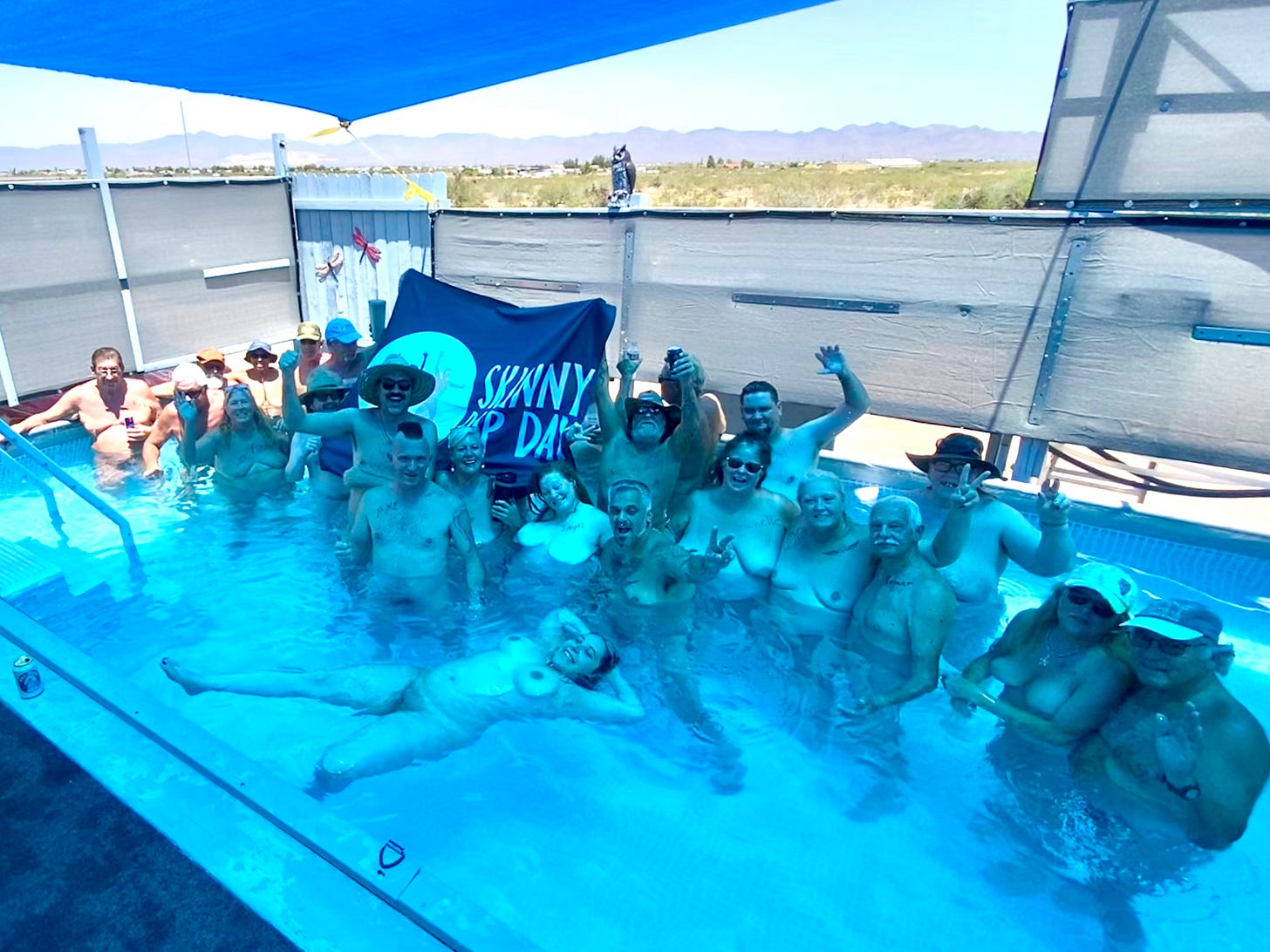 A group of smiling nude participants stands and floats in a clear blue pool at Golden Valley Sun Club in Arizona, celebrating Skinny Dip Day 2025. A “Skinny Dip Day” flag is held at the center, and the surrounding desert landscape is visible in the background under a bright blue sky. A group of smiling nude participants stands and floats in a clear blue pool at Golden Valley Sun Club in Arizona, celebrating Skinny Dip Day 2025. A “Skinny Dip Day” flag is held at the center, and the surrounding desert landscape is visible in the background under a bright blue sky.