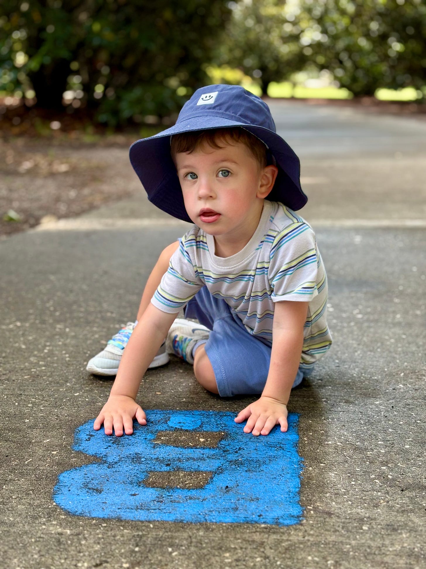Young child with a hat, blonde hair, and blue eyes drawing a b with chalk on the sidewalk Young child with a hat, blonde hair, and blue eyes drawing a b with chalk on the sidewalk