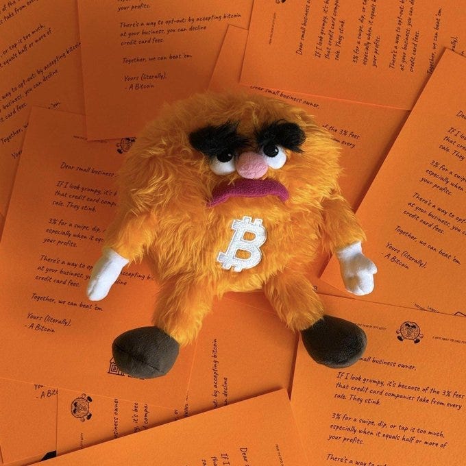 Orange furry plush toy shaped like the Bitcoin symbol with black eyebrows pink nose wide eyes and frowning mouth sits on a surface with white paws extended amid scattered orange paper letters containing text about credit offers business profiles debt collection and spam-like business propositions