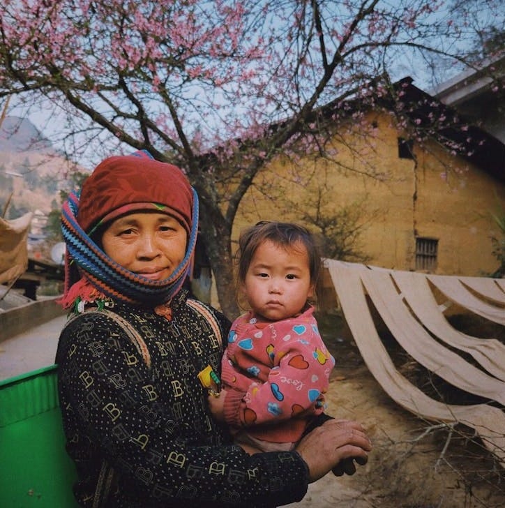 Free A mother and child stand in a rural village with blossoming trees, evoking a warm, cultural scene. Stock Photo