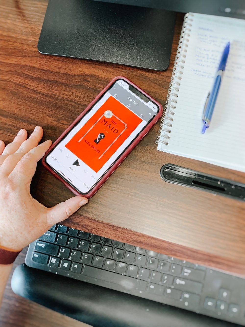 Picture of a cell phone on a desk next to a pen and notebook. The audiobook displayed on the phone is The Maid by Nita Prose.