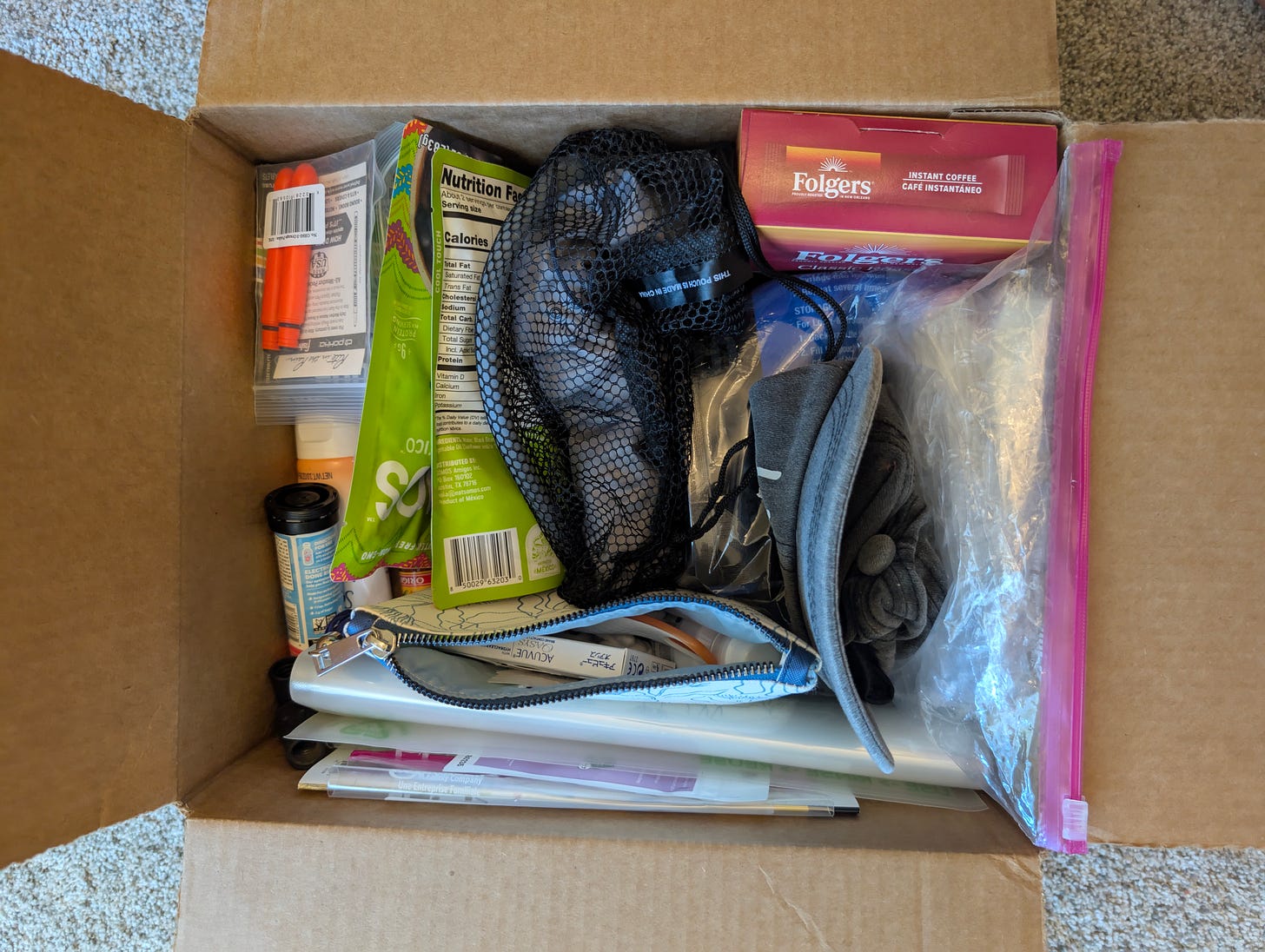 A cardboard resupply box filled with hiking essentials, including a mesh pouch, Folgers instant coffee packets, electrolyte tablets, a contact lens case, waterproof pens, and zip-top bags. Items are neatly organized and packed for an upcoming thru-hike.