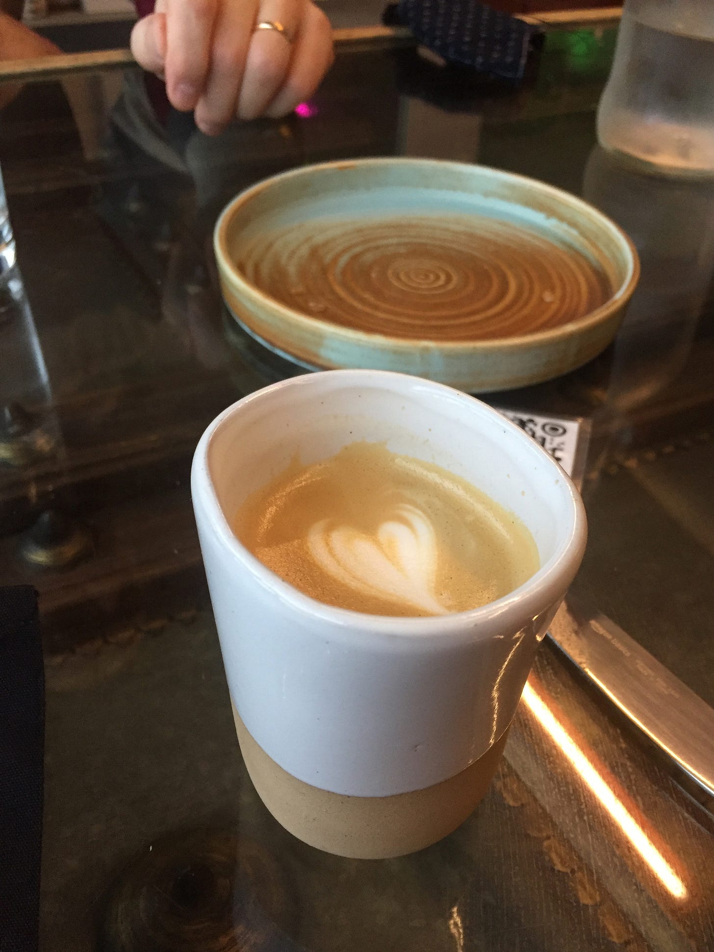 In the foreground, a white and tan handle-less mug of a cortado with a leaf drawn on top. Behind it, a teal green and light brown plate with walled sides sits empty. Jeff's hand is in the background behind it. In the foreground, a white and tan handle-less mug of a cortado with a leaf drawn on top. Behind it, a teal green and light brown plate with walled sides sits empty. Jeff's hand is in the background behind it.