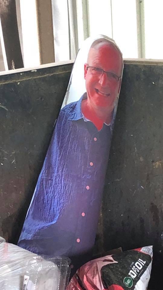 A tall, almost cylindrical inflated thing with a photograph of a man printed on it. He's middle-aged, smiley, and wearing a blue button-up shirt. He's propped up in the corner of a dumpster, among other trash.