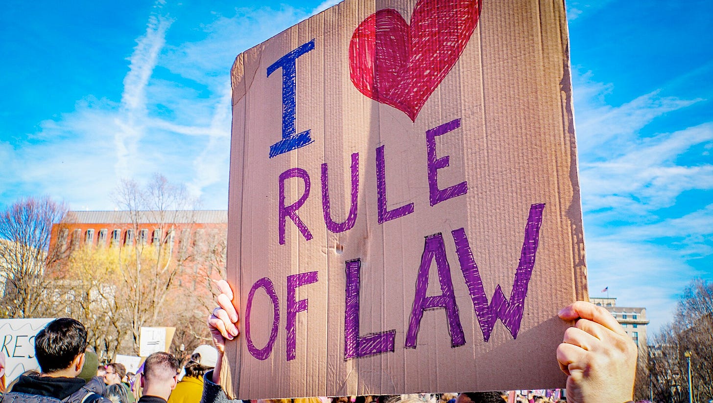 with a blue sky and official building in the background a handmade brown cardboard sign reads I heart rule of law with a blue sky and official building in the background a handmade brown cardboard sign reads I heart rule of law