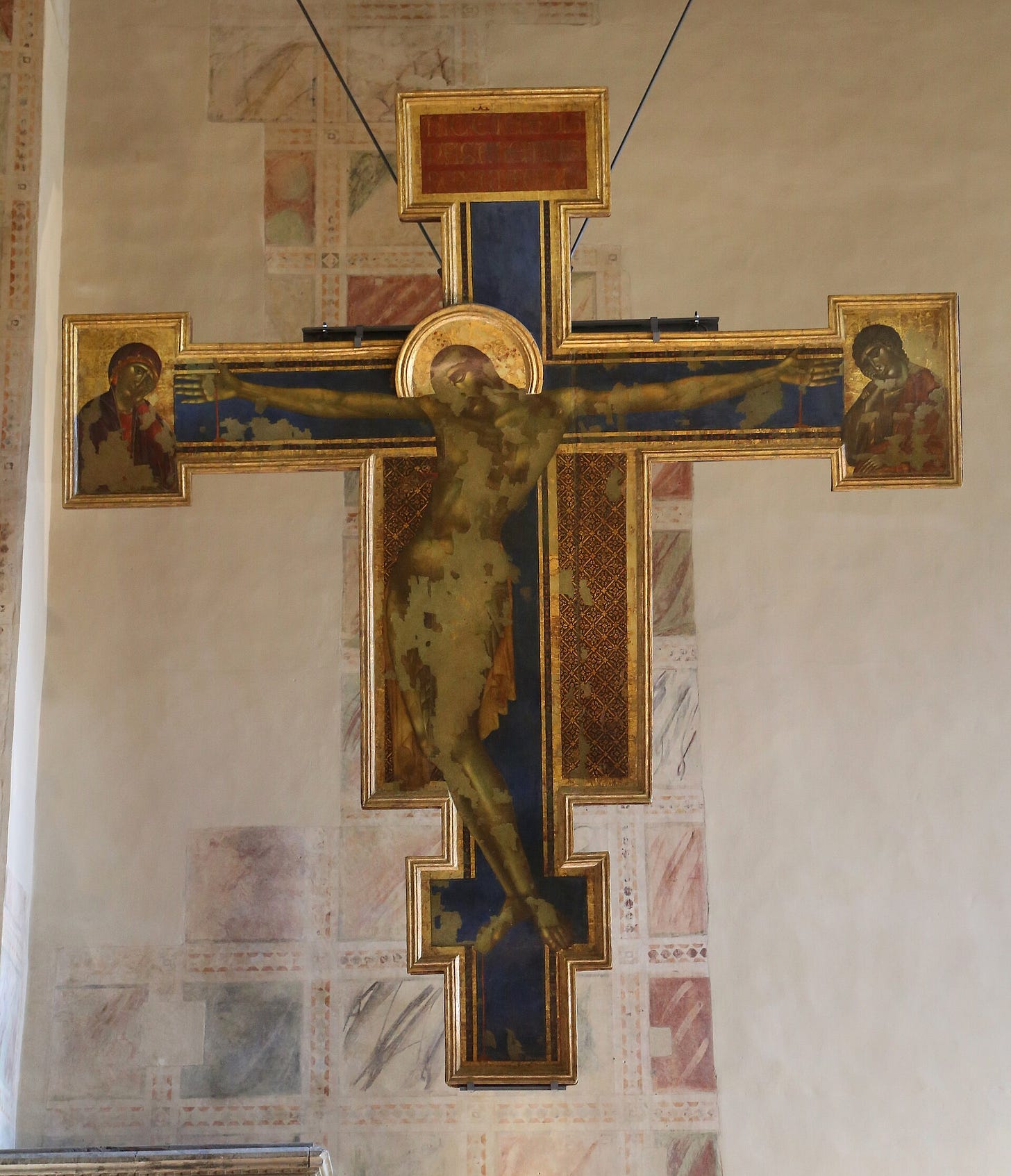A tall painted wooden cross on a gold ground: Christ hangs with his body curved to one side and his head bowed, with small half-length figures of the Virgin and Saint John at the ends of the arms, large areas of the paint are worn away. A tall painted wooden cross on a gold ground: Christ hangs with his body curved to one side and his head bowed, with small half-length figures of the Virgin and Saint John at the ends of the arms, large areas of the paint are worn away.
