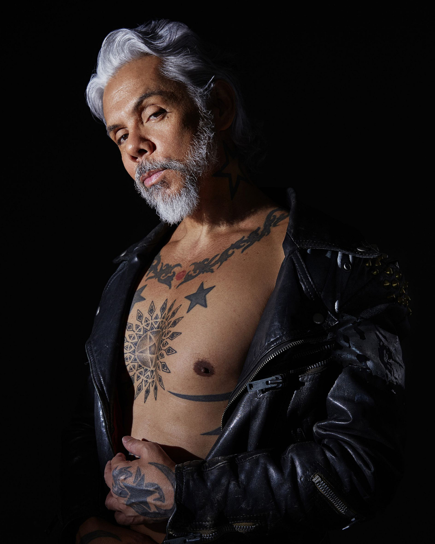 Older Blasian person with gray hair and a beard and mustache poses confidently against a dark black background. They wear a studded leather jacket, revealing intricate chest tattoos. They are looking confidently into the camera, and have their left hand resting on their torso. Older Blasian person with gray hair and a beard and mustache poses confidently against a dark black background. They wear a studded leather jacket, revealing intricate chest tattoos. They are looking confidently into the camera, and have their left hand resting on their torso.