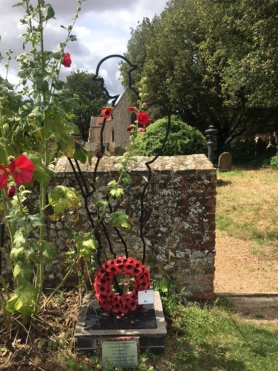 Image of a Tommy memorial which is a black metal outline of a soldier holding a gun, erected in front of a stone church wall, with a poppy wreath in foreground, and hollyhocks in bloom. Church just visible in background