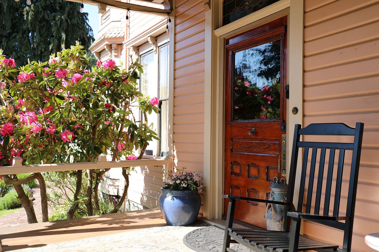 The front door of the Adams Pragge House from the covered porch, with a rocking chair at right and a pink rhododendron blooming at left.