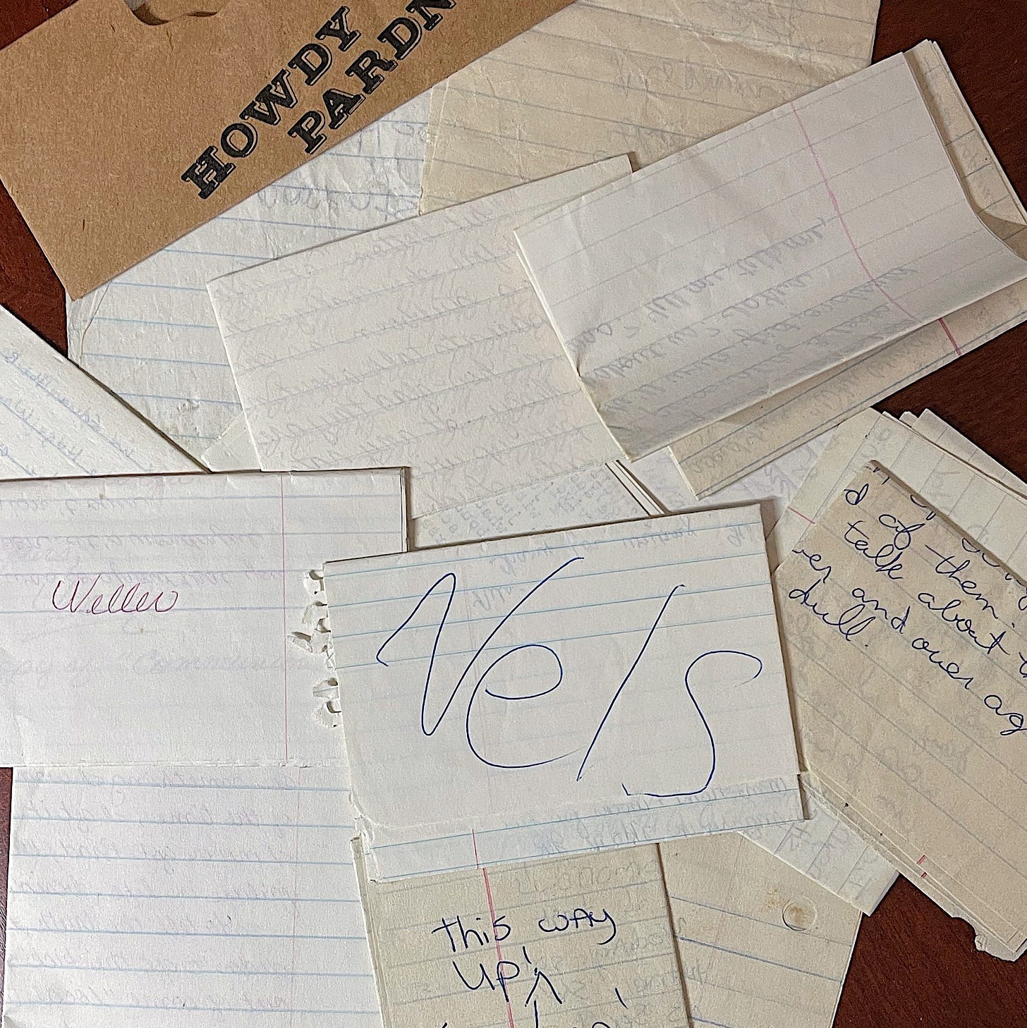 A scattered collection of old, handwritten notes on torn pieces of lined notebook paper, some yellowed with age. Visible words include “Neller,” “Nels,” and “this way up!” written in different handwriting styles and ink colors. A Kraft paper tag reading "Howdy Pardner" peeks out at the top. The notes overlap on a reddish wooden surface, evoking a sense of nostalgia and memory.