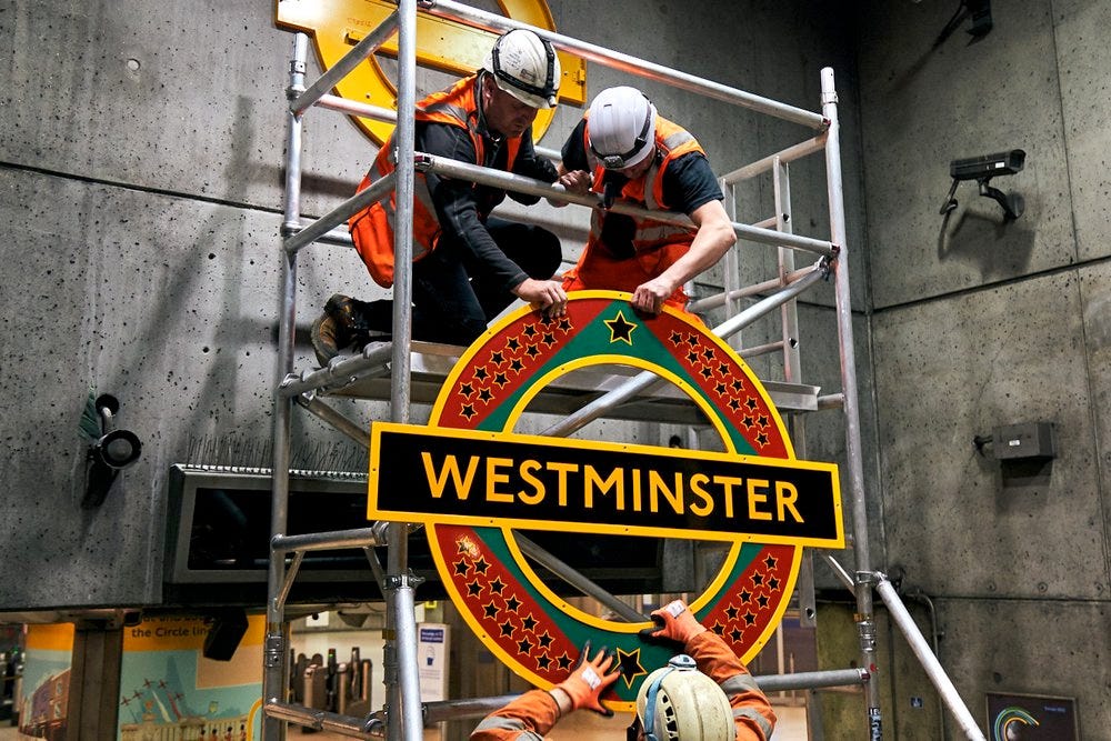 Three workmen in a metal scaffold erect a colourful red, green and yellow London Underground roundel designed by the artist Larry Achiampong Three workmen in a metal scaffold erect a colourful red, green and yellow London Underground roundel designed by the artist Larry Achiampong