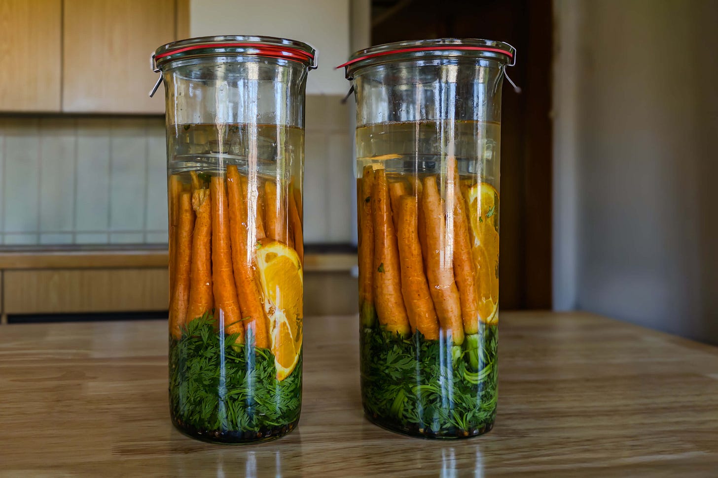 Zwei hohe Weck-Gläser mit Karotten und Karottengrün, geschichtet mit Orangenscheiben und Gewürzen, mit Lake aufgegossen und verschlossen auf einem Holztisch in der Küche. Oben im Glas sind die Glasgewichte erkennbar, welche die Karotten unter die Salzlake drücken.