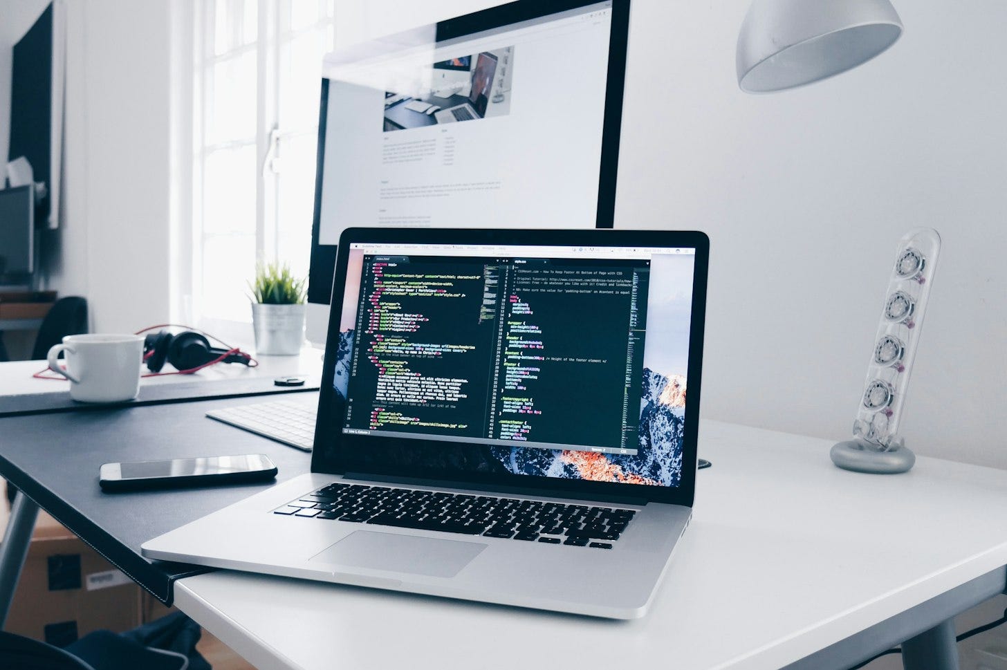 An open laptop showing lines of code in a dark editor window on a white desk, with an external monitor, a coffee cup and a phone nearby. An open laptop showing lines of code in a dark editor window on a white desk, with an external monitor, a coffee cup and a phone nearby.