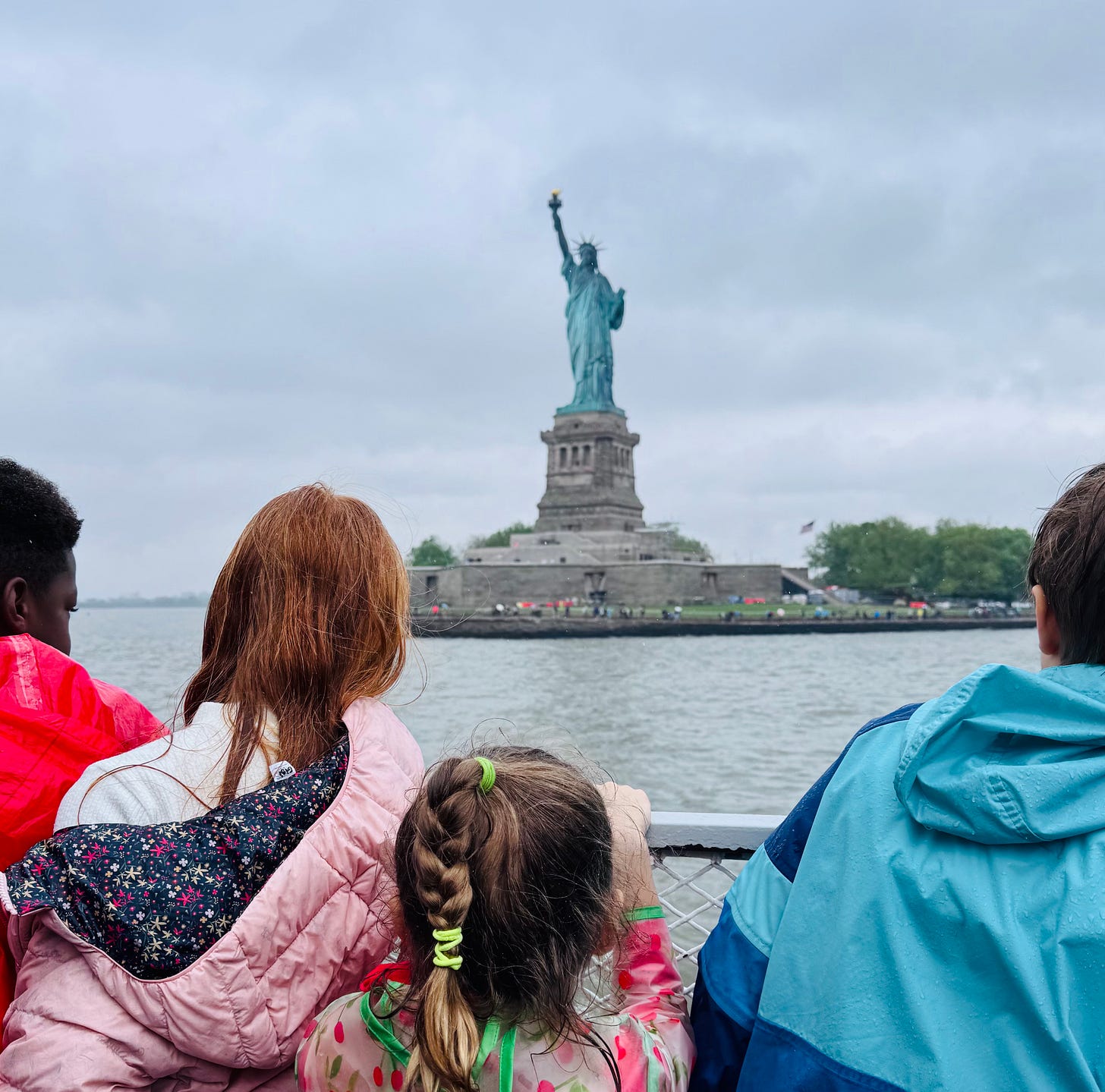 Children stand with their backs facing the camera. They look over a fence at the Statue of Liberty. It's overcast and raining.