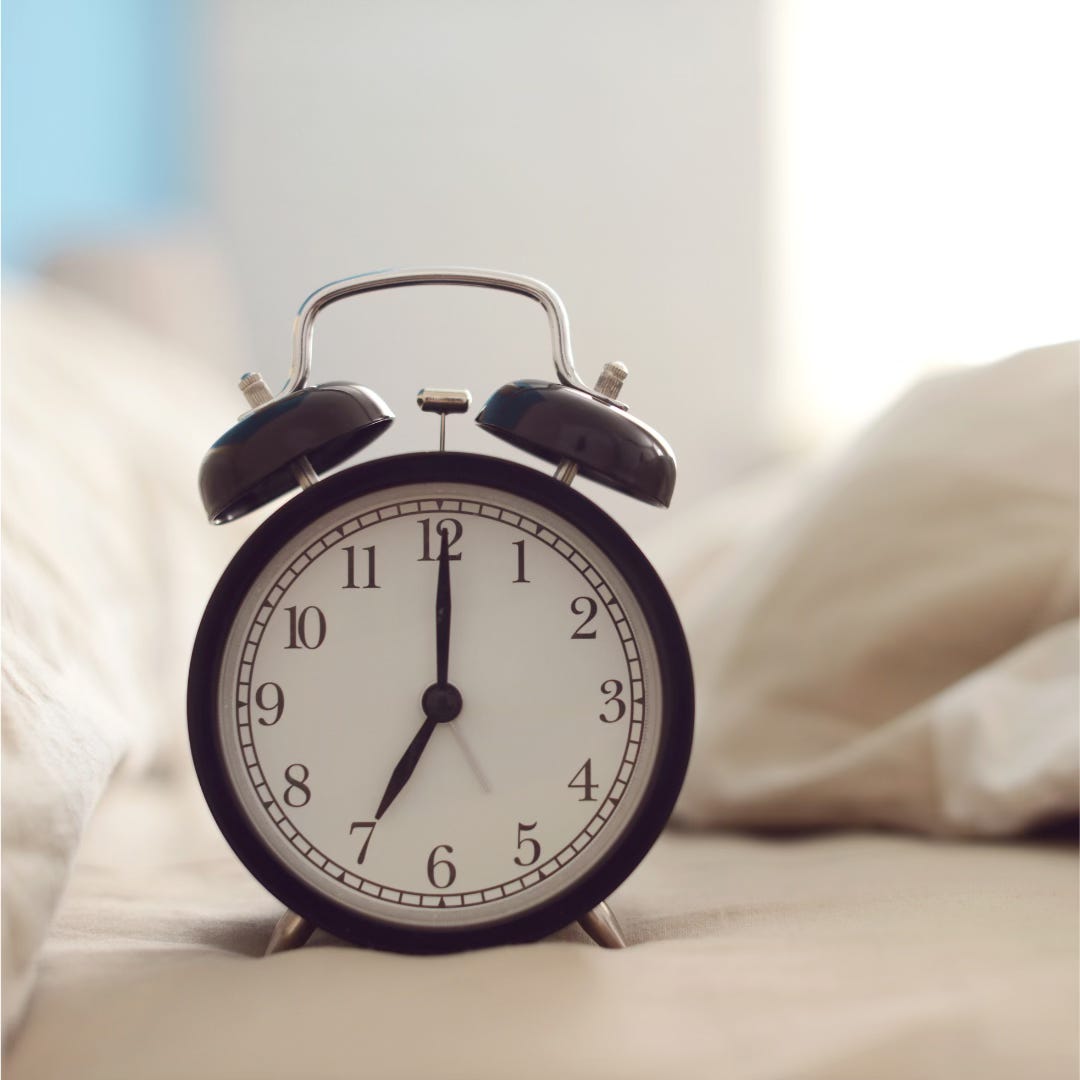 A photo of an alarm clock sitting on a bed made with cream coloured sheets.