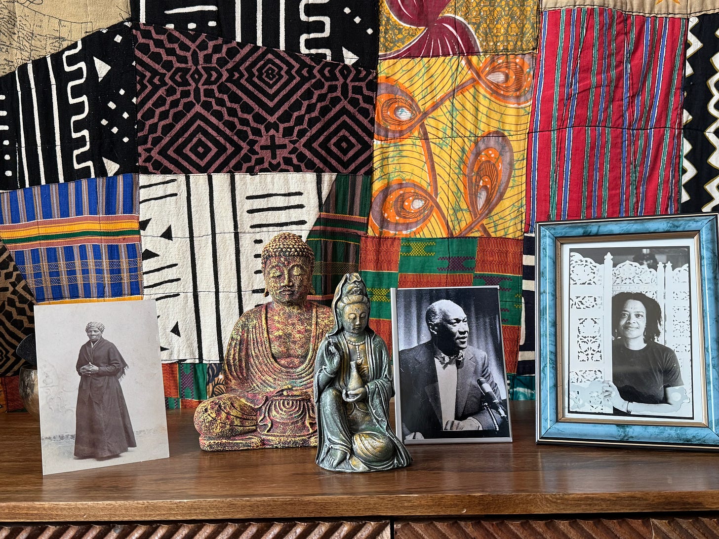 A mudcloth quilt forms the backdrop. In the foreground, from left to right: a photograph of Harriet Tubman, a Buddha statue, a Kuan Yin statue, a photograph of Rev. Dr. Leon Wright, and a photograph of Toni Cade Bambara.