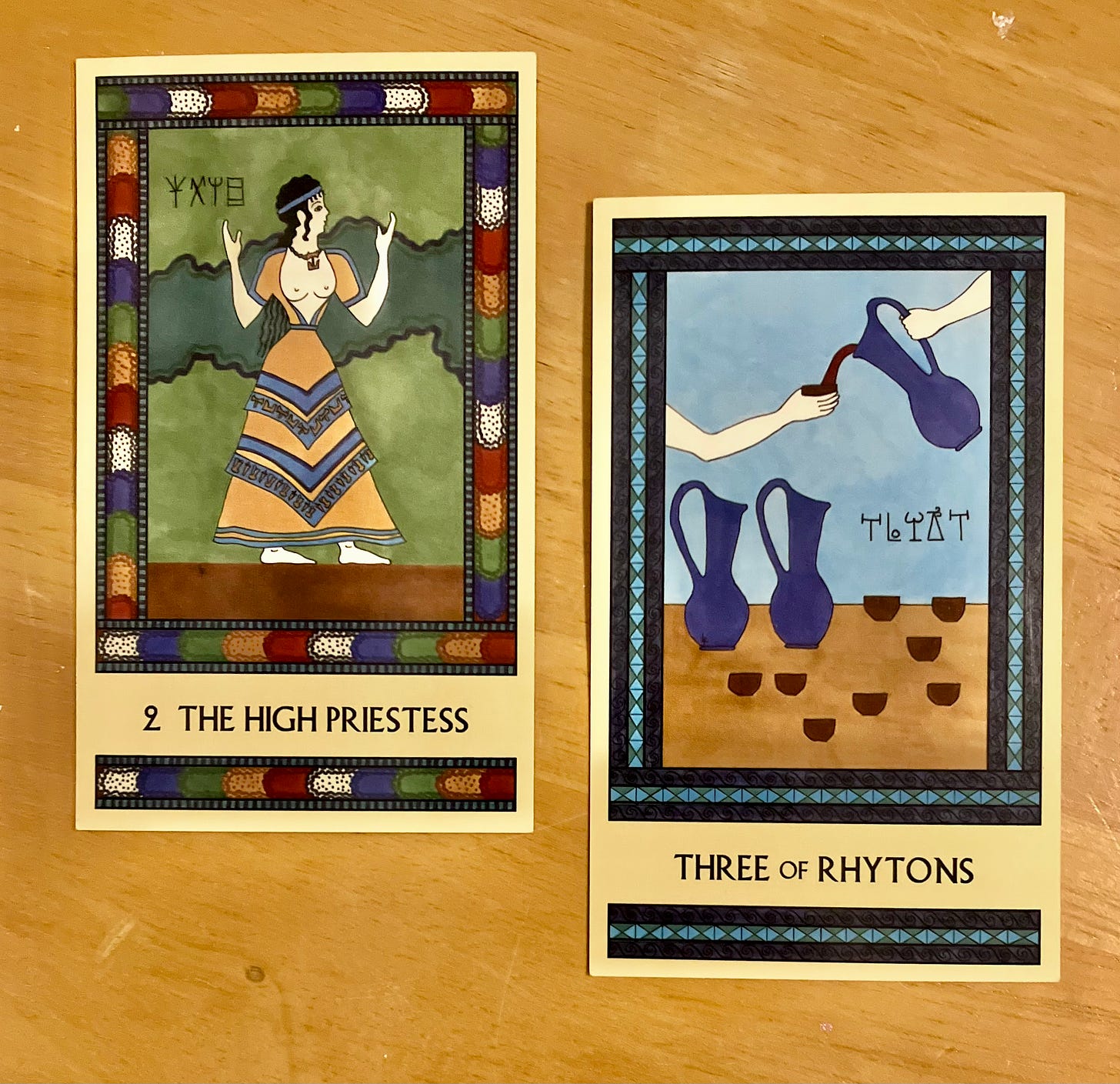 Two Minoan Tarot cards side by side on a golden wood surface. The High Priestess has a bright multicolor border. It shows a Minoan priestess standing, holding her hands up in a sacred gesture. She is facing right. The Three of Rhytons is painted in shades of blue and tan. It shows a hand holding a blue pitcher, pouring red wine from that pitcher into a cup held by another hand. Also pictured are two more blue pitchers and several more cups. 