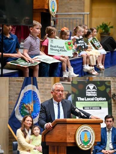 Children with Real Food Strong Kids signs above Kennedy speaking beside a The Local Option poster