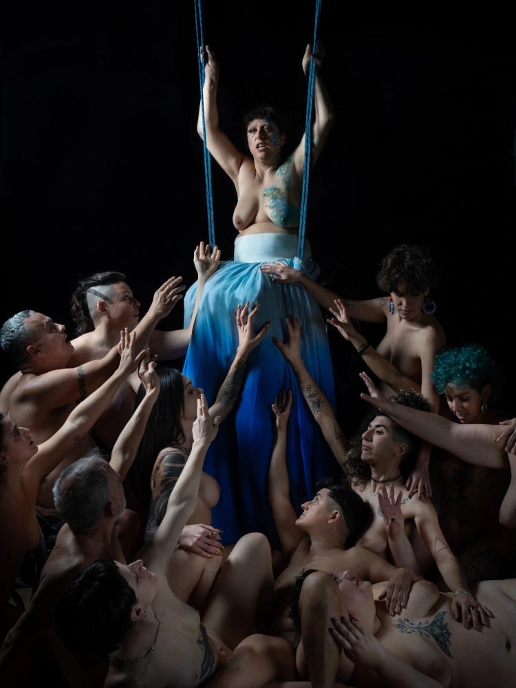 Un gruppo di persone nude, dai corpi diversi per genere, età e caratteristiche fisiche, si accalca in basso, rivolgendosi con le mani alzate verso una figura centrale sospesa su corde azzurre. La persona al centro, con il corpo parzialmente coperto da una lunga gonna sfumata blu, guarda in alto mentre si tiene alle corde, come su un trapezio. L'atmosfera teatrale, il fondo nero e la composizione piramidale richiamano un’estetica classica e sacra, trasformata qui in una potente immagine di desiderio, alleanza e celebrazione delle soggettività crip-queer.