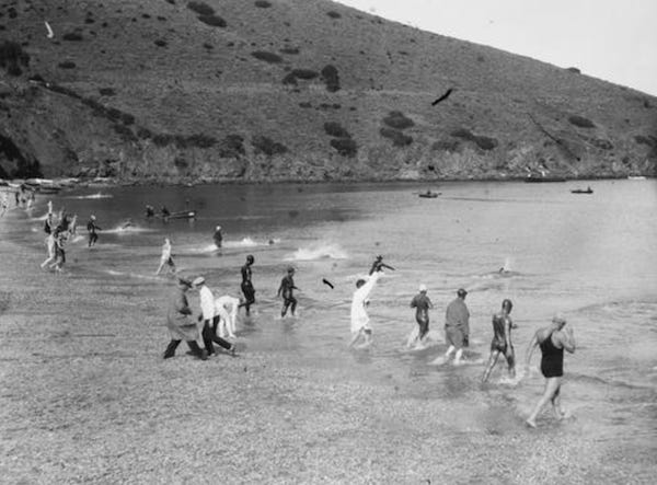A black and white photo of a couple dozen people in varying states of dress wading into the water. A black and white photo of a couple dozen people in varying states of dress wading into the water.