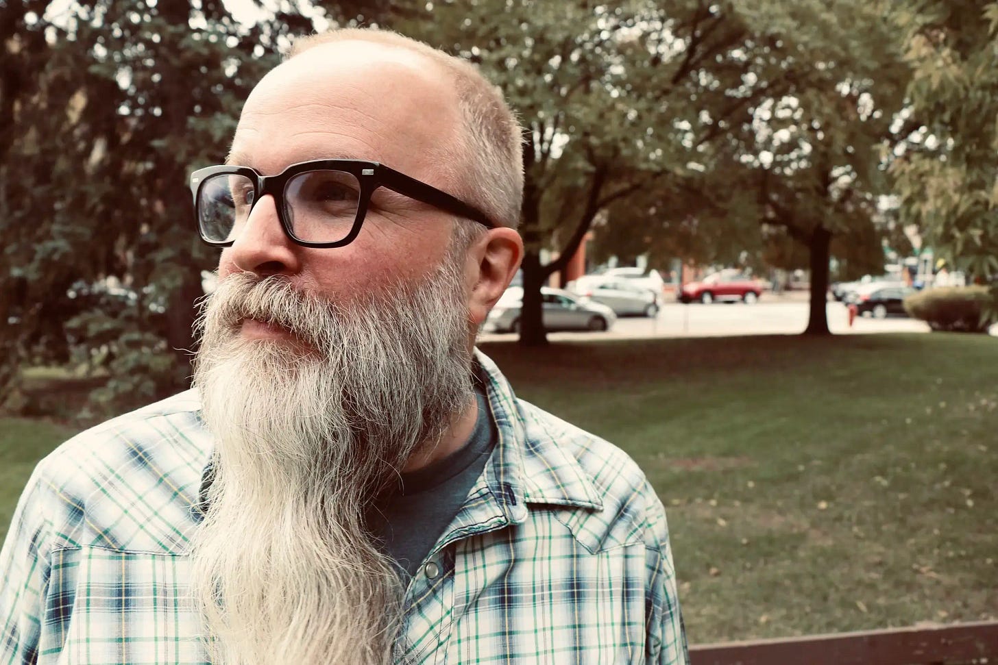 A middle-aged man with a full, long gray beard and receding hairline sits outdoors wearing black-rimmed glasses and a blue and green plaid button-up shirt. He's looking off to the side with a contemplative expression. Behind him is a park-like setting with large trees creating dappled sunlight, and several cars are visible parked in the background. The lighting has a warm, golden quality suggesting late afternoon or early evening. A middle-aged man with a full, long gray beard and receding hairline sits outdoors wearing black-rimmed glasses and a blue and green plaid button-up shirt. He's looking off to the side with a contemplative expression. Behind him is a park-like setting with large trees creating dappled sunlight, and several cars are visible parked in the background. The lighting has a warm, golden quality suggesting late afternoon or early evening.