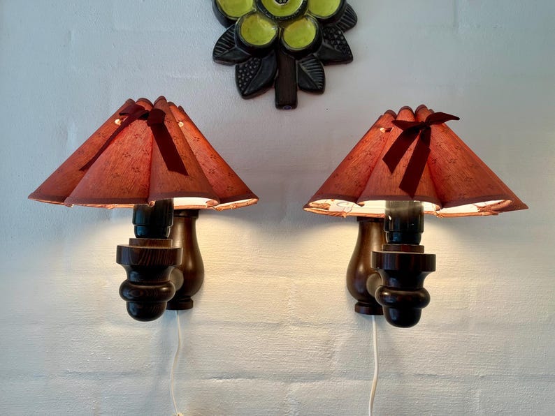 May include: Two vintage wall sconces with dark brown wooden bases and reddish-brown lampshades. The lampshades have a unique, scalloped design and are adorned with a dark brown bow. A decorative wall hanging is above the sconces. May include: Two vintage wall sconces with dark brown wooden bases and reddish-brown lampshades. The lampshades have a unique, scalloped design and are adorned with a dark brown bow. A decorative wall hanging is above the sconces.