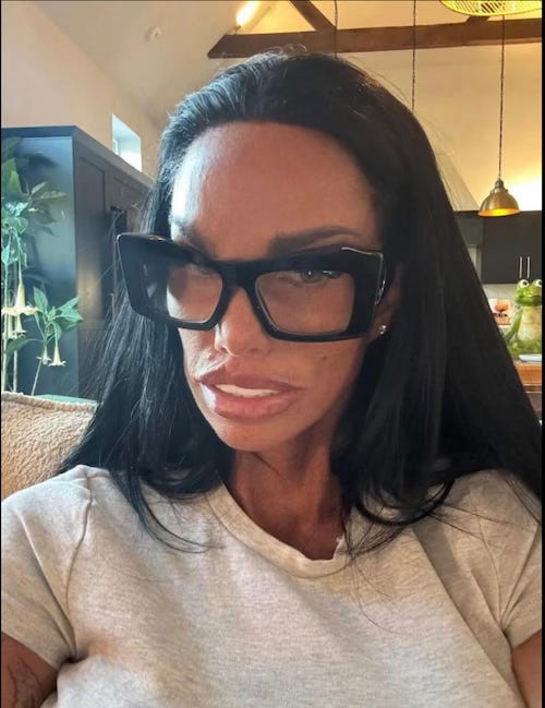 Katie Price in larger thick-framed glasses, with dark hair, her trademark tan and very white teeth. Numerous surgical enhancements have left her looking very taut and slim in the face.