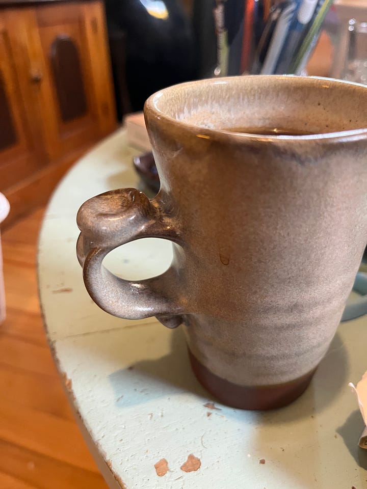 two pottery mugs, one blue and squat, one gray and skinny. Both have a dot of clay at the top of the handle upon which to rest your thumb