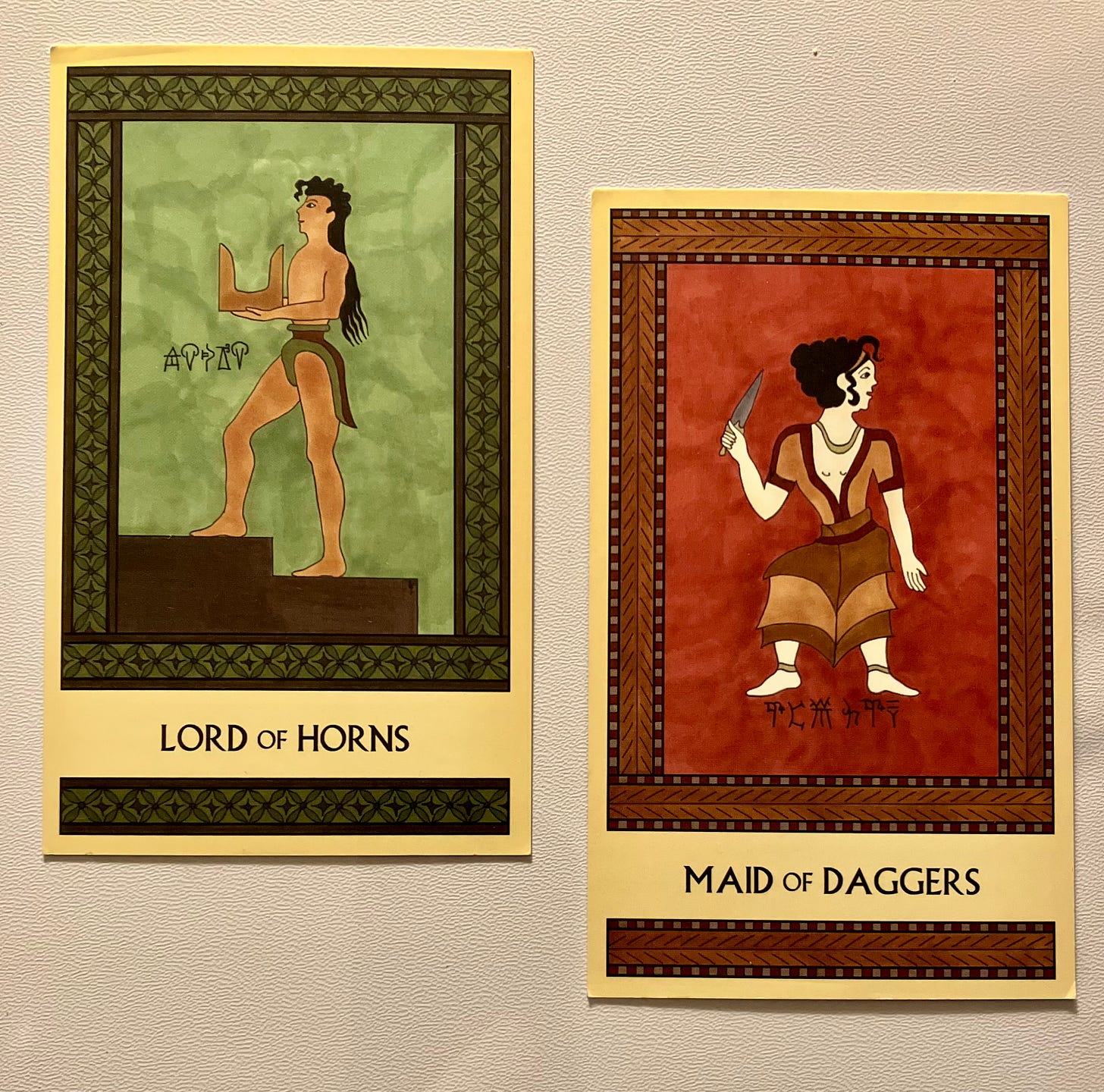 Two Minoan Tarot cards side by side on a white surface. The Lord of Horns card, on the left, is painted in shades of deep green and brown. It shows a Minoan man with tan skin and long hair, wearing a loincloth and codpiece. He is facing left, walking up a flight of steps, and holding a large pair of sacred horns. The Maid of Daggers card, on the right, is painted in shades of deep red and tan. It shows a light-skinned Minoan girl wearing a dress, a necklace, and anklets. She is standing, facing forward but with her head turned to the right. In her right hand she holds up a dagger. 