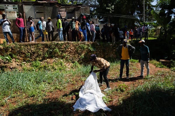 People stand outside on a stone wall as a person below holds a white sheet shrouding a body on ground with sparse grass.