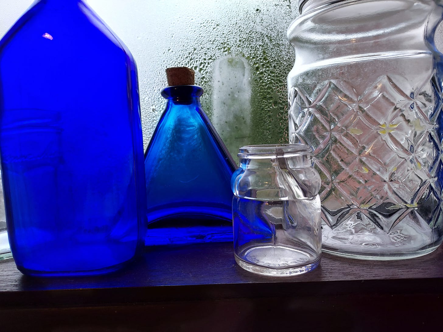 Several glass containers sit on a sill in front of a fogged up window. On the far left are two deep blue glass containers, one tall and syllindrical tapering to a narrow mouth, the other triangular in shape with a cork stopper. In the center right of the frame is a small transparent and empty glass inkwell. On the far right of the frame, taking up about a third of the picture area, is a large clear glass jar with a diamond pattern on its surface. Several glass containers sit on a sill in front of a fogged up window. On the far left are two deep blue glass containers, one tall and syllindrical tapering to a narrow mouth, the other triangular in shape with a cork stopper. In the center right of the frame is a small transparent and empty glass inkwell. On the far right of the frame, taking up about a third of the picture area, is a large clear glass jar with a diamond pattern on its surface.