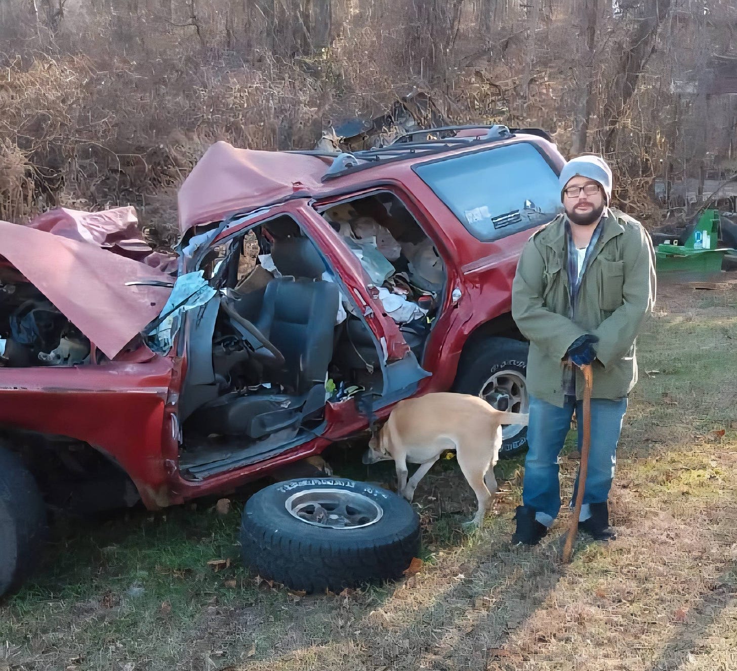 A man leaning on a cane standing next to the wreckage of a red Toyota 4Runner, with a dog sniffing the vehicle.