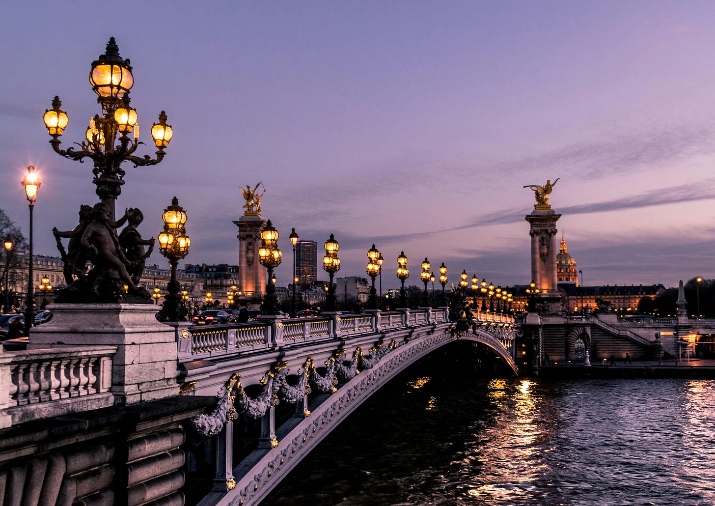 An ornate bridge over the River Seine in Paris at dusk. All of the lamps are lit, and the river reflects the light.
