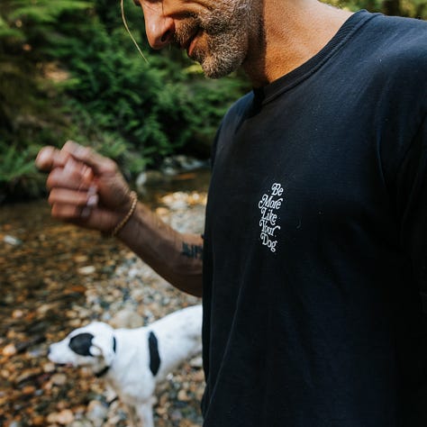 awkward man frolics around stream with dogs wearing very nice merch from be more like your dog