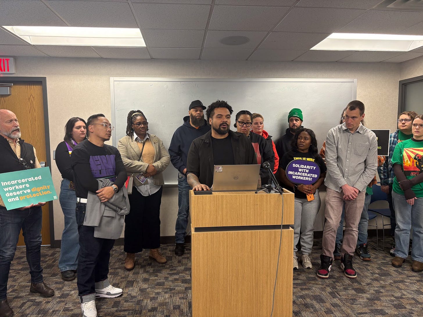 a dozen people stand behind a Black man wearing a black shirt speaking at a podium