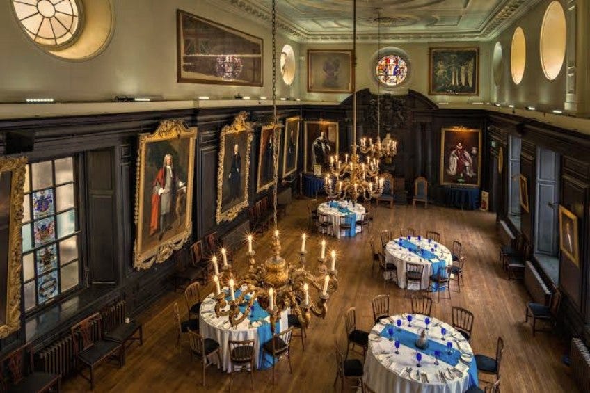 An elevated, wide-angle view of the historic Great Hall inside Apothecaries' Hall, London. The room features dark, ornate 17th-century oak paneling lined with large framed oil portraits, massive hanging brass chandeliers, and several round dining tables meticulously dressed in crisp white and vibrant blue linens for a formal event. An elevated, wide-angle view of the historic Great Hall inside Apothecaries' Hall, London. The room features dark, ornate 17th-century oak paneling lined with large framed oil portraits, massive hanging brass chandeliers, and several round dining tables meticulously dressed in crisp white and vibrant blue linens for a formal event.