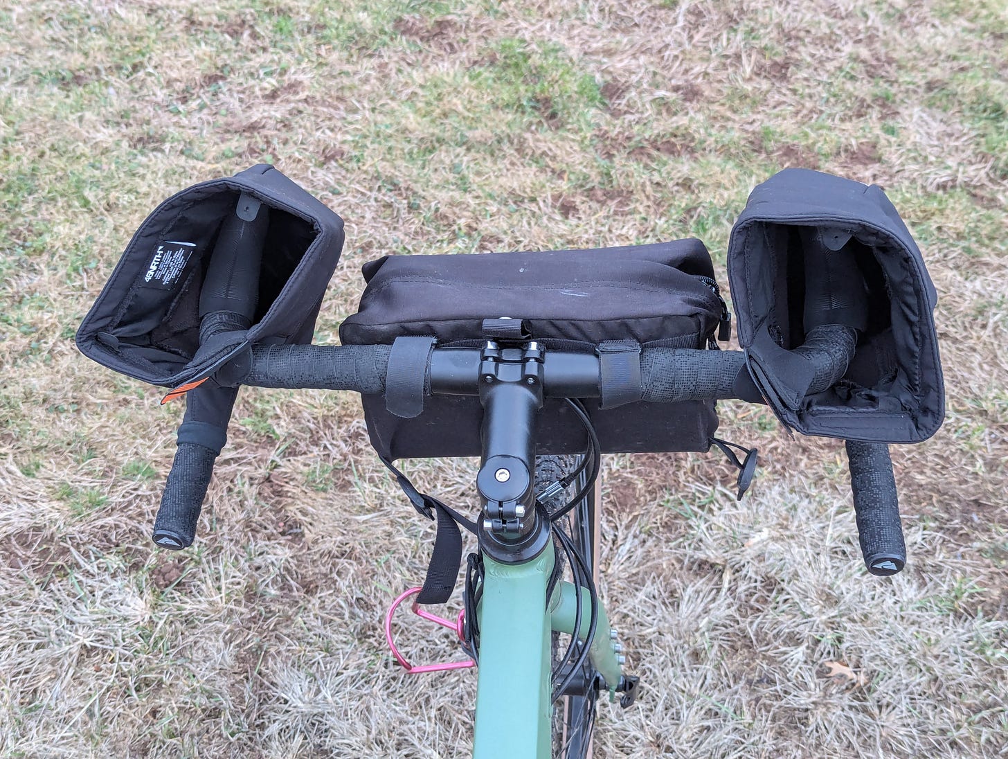 Photo of a bicycle, looking down as if seated on the bike. The bike is green with curved drop bars with black tape. The "hoods" where the brakes and shifters are positioned are surrounded by soft, pliable frabric pogies. The rider sticks their hands in the pogies and then grips the levers. There is also a small black handlebar bag velcroed to the handlebar.