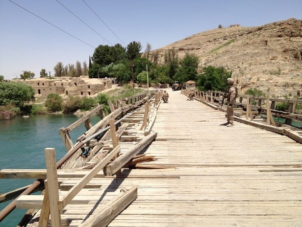 This image of a bridge taken from one end looking along its length is in Kajaki, Afghanistan (north central). A group of Marines in military gear crossing a damaged wooden bridge over a body of water in a dry, mountainous region. The bridge has broken and missing planks, along with makeshift wooden railings, indicating wear or conflict-related damage. In the background, there are mud-brick buildings, green vegetation, and hills. One Marine is standing guard on the right side of the bridge midway across, holding a rifle, while others are walking further ahead. The sky is clear and bright during a sunny day. This image of a bridge taken from one end looking along its length is in Kajaki, Afghanistan (north central). A group of Marines in military gear crossing a damaged wooden bridge over a body of water in a dry, mountainous region. The bridge has broken and missing planks, along with makeshift wooden railings, indicating wear or conflict-related damage. In the background, there are mud-brick buildings, green vegetation, and hills. One Marine is standing guard on the right side of the bridge midway across, holding a rifle, while others are walking further ahead. The sky is clear and bright during a sunny day.