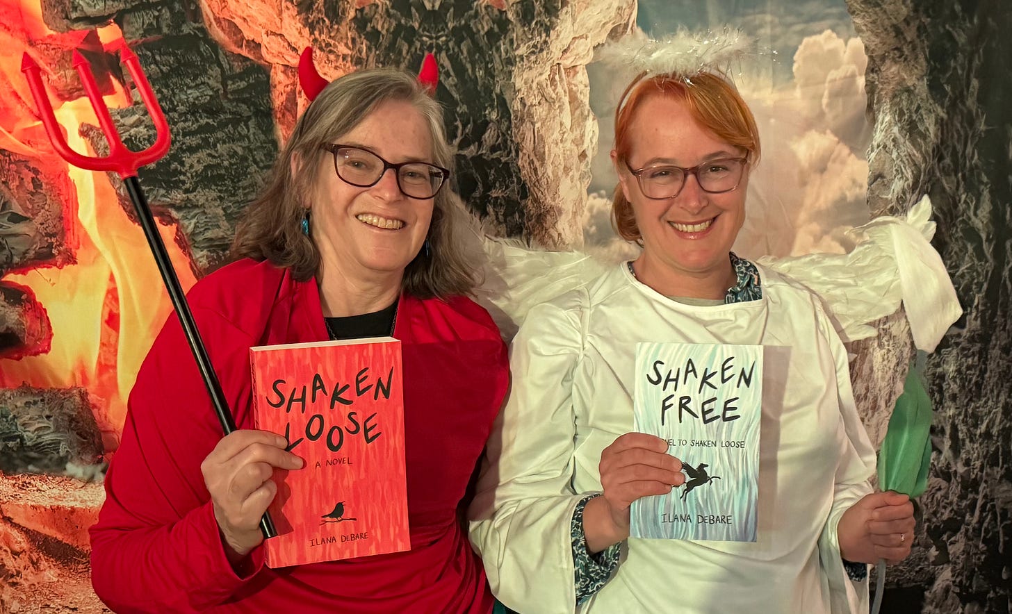 Two women in devil and angel costumes in front of a backdrop of Hellish flames and Heavenly lights and clouds, each holding one of Ilana's books