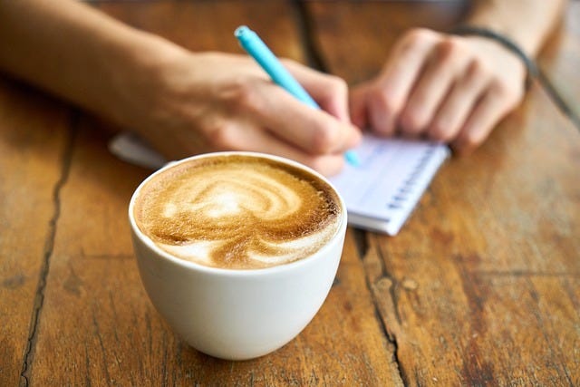 Photo of a frothy cup of cappuccino on top of a wooden table. In the background is a hand holding pen to a small notebook.