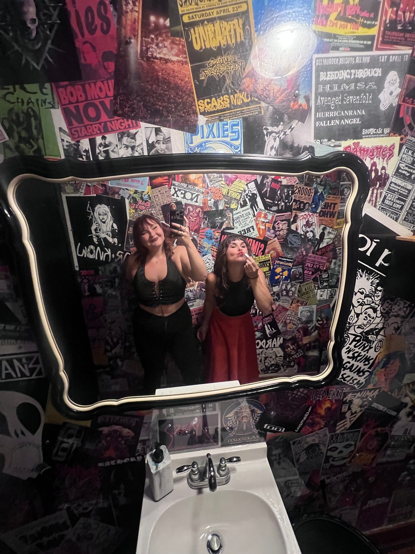 Two beautiful cunty women stand in front of a bathroom sink. The bathroom walls are papered in fliers from punk shows. 