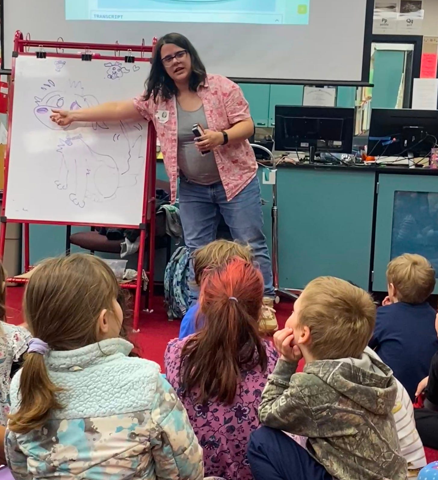 Lindsay Hornsby, a dark haired cartoonist with glasses, gestures to a whiteboard with a worried puppy drawn on it. She's speaking about how to show character emotions to several elementary kids, seen from behind.