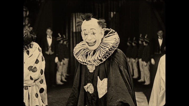 Chaney smiling in costume: Lon Chaney as the tragic clown ‘HE,’ smiling in full ruffled costume, surrounded by circus performers in He Who Gets Slapped (1924).