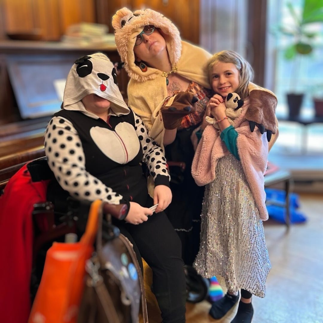 Three people in whimsical animal-themed costumes pose together indoors. One person, seated on a walker/mobility device, wears a black-and-white spotted hoodie with a panda face on the hood. Another, standing behind them, wears a fluffy beige sloth costume with a hood, extending one of the costume's sloth arms. A young child in a sparkling dress and a pink cape with a small plush panda in their arms smiles brightly.