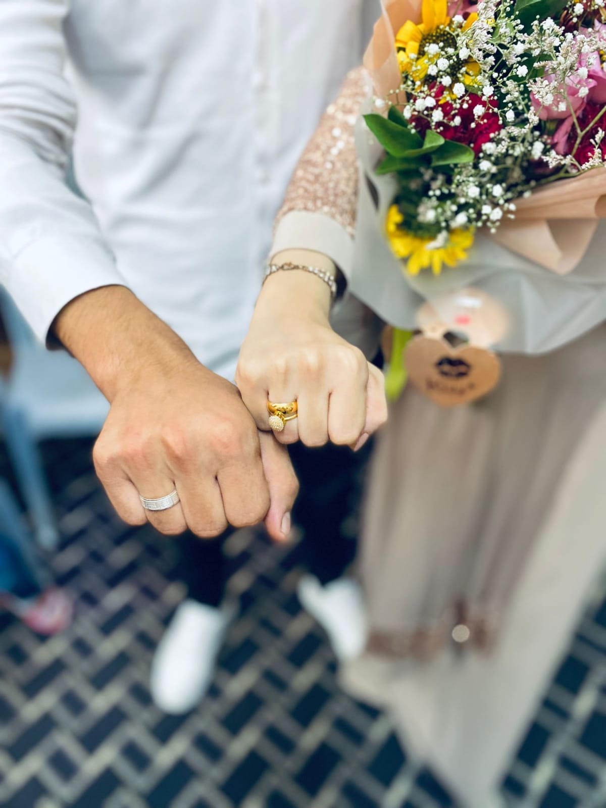 A close-up, top-down view of two people standing side by side, extending their hands forward to show matching rings on their ring fingers. One hand wears a simple silver band, the other a gold ring with a decorative setting. To the right, one person holds a bouquet of colorful flowers wrapped in light paper. The background shows patterned paving stones and softly blurred clothing and shoes. A close-up, top-down view of two people standing side by side, extending their hands forward to show matching rings on their ring fingers. One hand wears a simple silver band, the other a gold ring with a decorative setting. To the right, one person holds a bouquet of colorful flowers wrapped in light paper. The background shows patterned paving stones and softly blurred clothing and shoes.