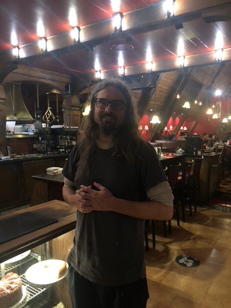 Jess Coble aka The Zero Collective stands in side a brightly lit, and architecturally strange los angeles valley deli. They are smiling, with black rim glasses a goateee and long gorgeous curly hair. 