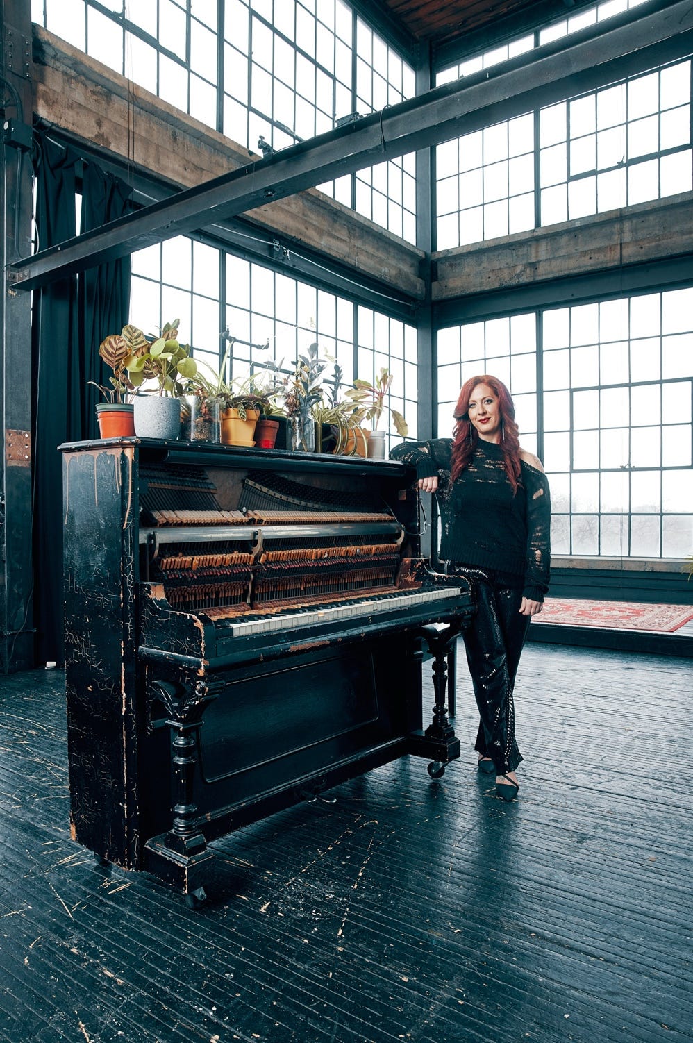 Cheryl Duvall stands in a warehouse-like space next to an upright piano. The room is lit from floor-to-ceiling windows. She is wearing a loose, black, off the shoulder jumper; the piano is also black and looks well-used. There is a row of houseplants on its top.