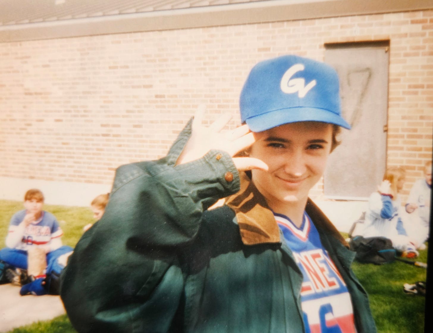 An old photograph of Emily at age 15, wearing the school softball uniform and hat, with a baggy green coat over top. Emily is smiling slightly into the camera and giving an awkward wave.
